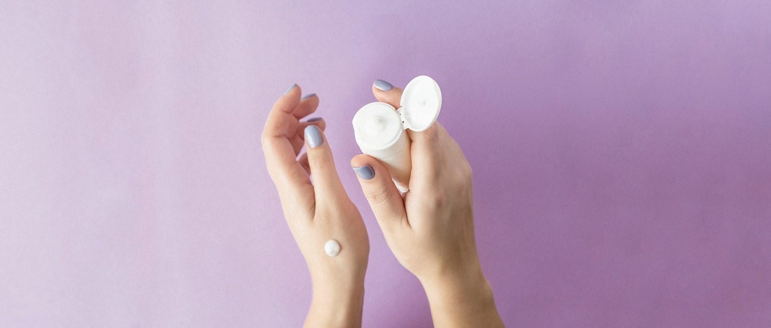 Manos con esmalte de uñas azul pálido sosteniendo un envase blanco para el cuidado de la piel con crema en una mano sobre fondo morado.
