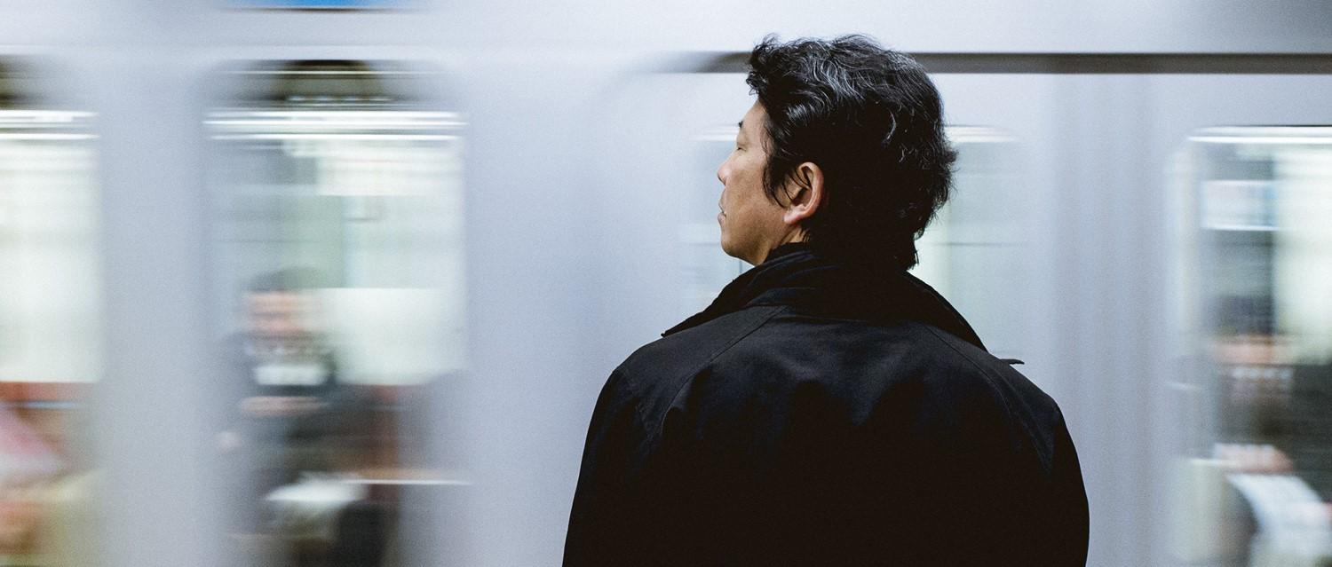 Persona con abrigo negro esperando en el andén del metro con un tren borroso pasando al fondo.