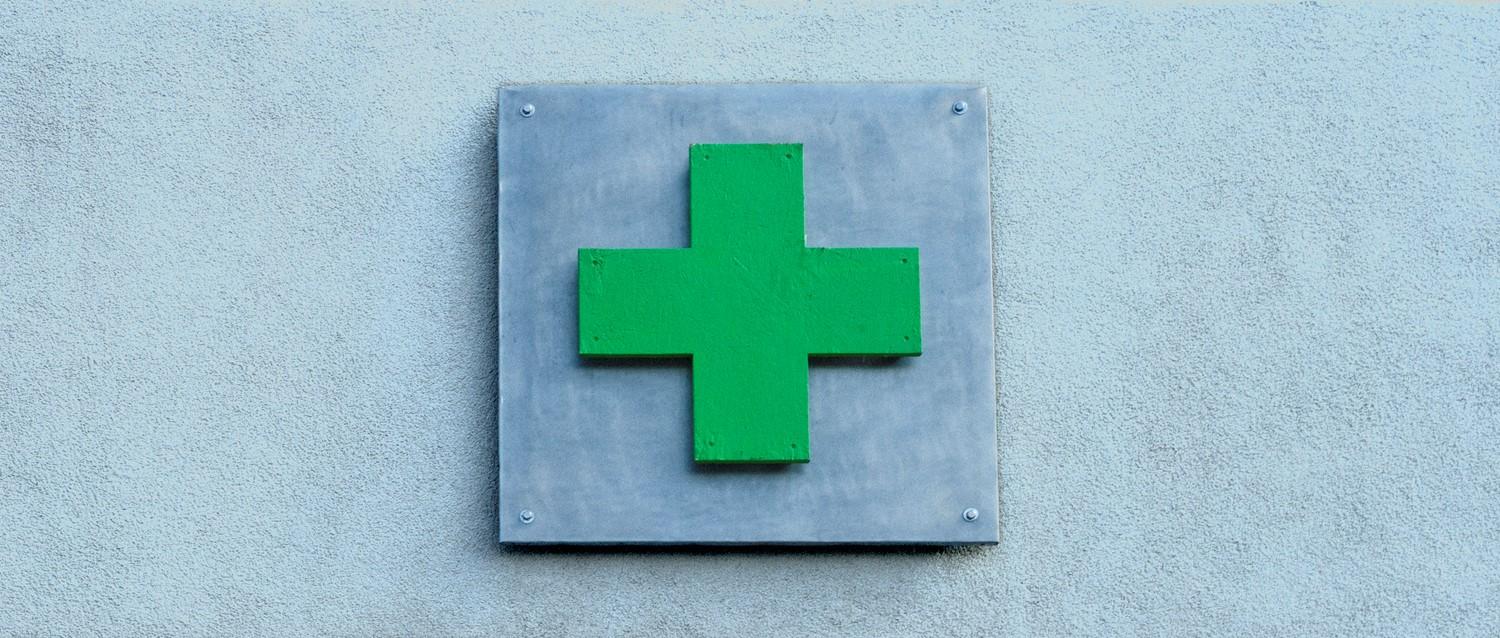 Green medical cross sign mounted on a grey metal plate against a light blue wall, indicating a healthcare facility.
