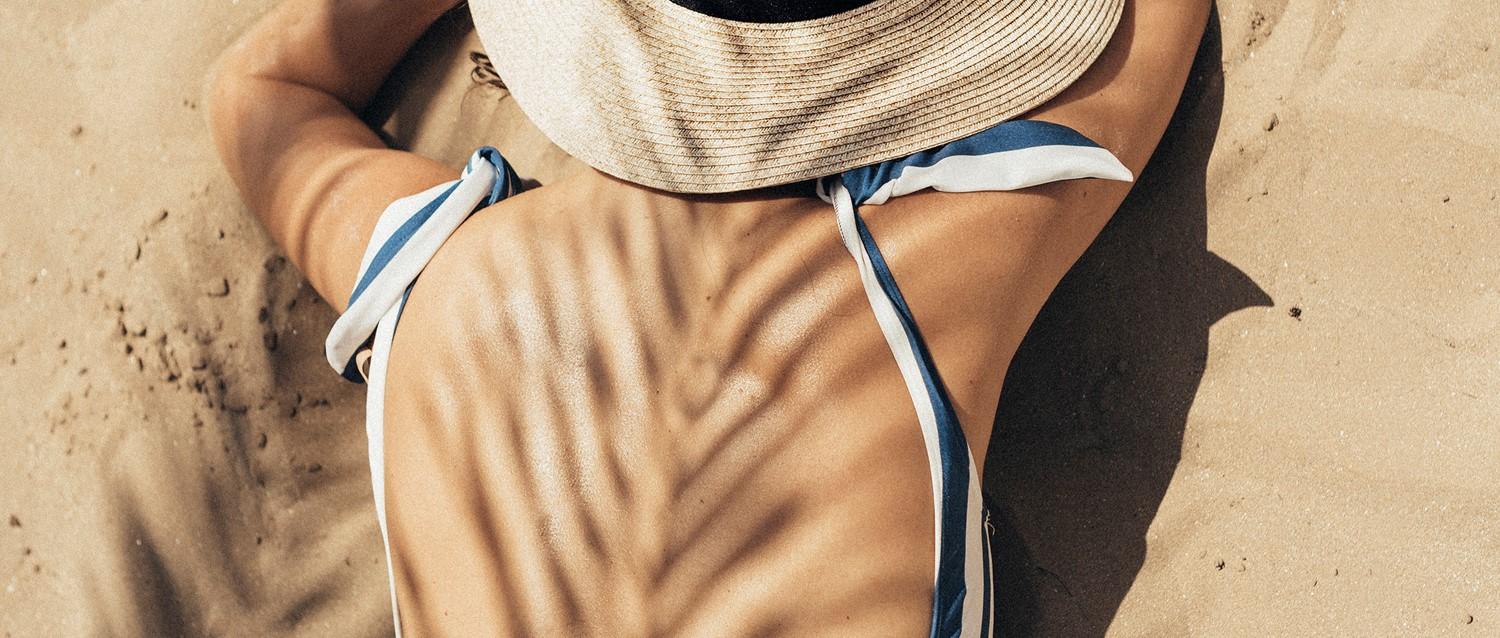 Persona relajándose en la arena de la playa con sombrero de paja y bikini blanco y azul, con la sombra de una palmera sobre su cuerpo.